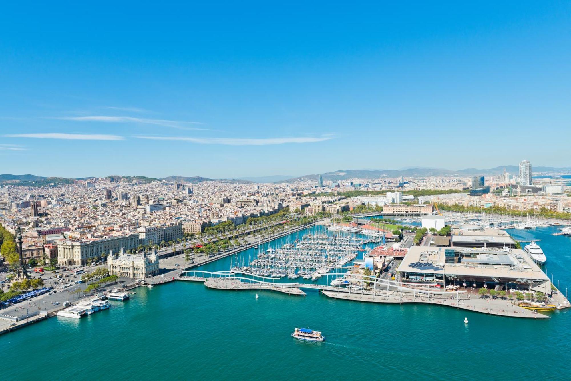 Eurostars Grand Marina Hotel Gl Barcelona Exterior photo The photo depicts a coastal city view, likely of a vibrant harbor or marina. In the foreground, there is a clear blue body of water with a few small boats. The harbor features numerous yachts and boats docked in the marina. 

Surrounding the water ar