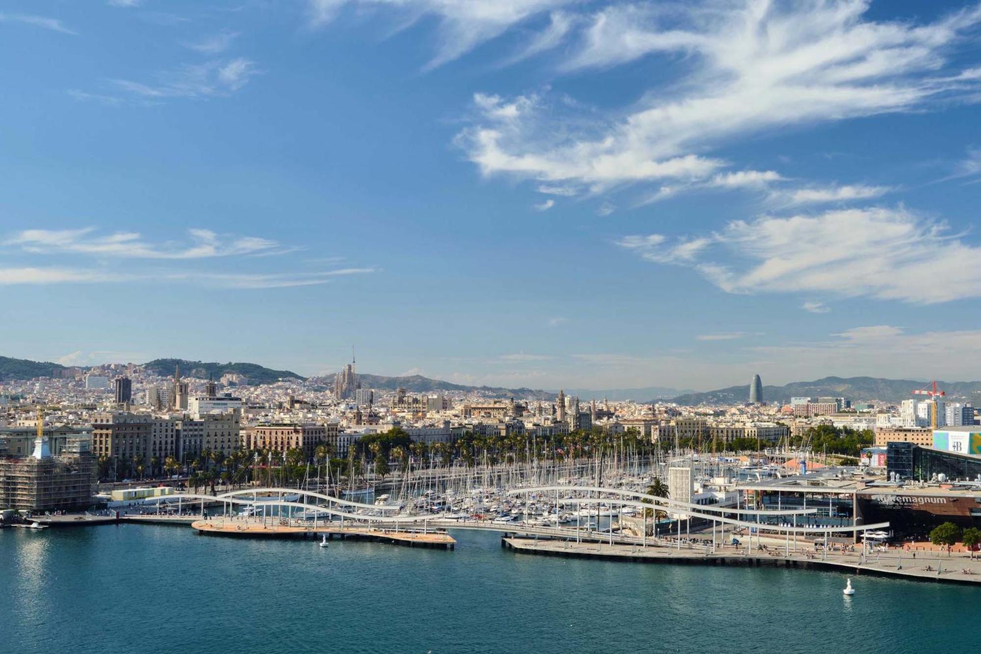 Eurostars Grand Marina Hotel Gl Barcelona Exterior photo The photo shows a scenic view of a coastal city, likely Barcelona, with a harbor area prominent in the foreground. There are several boats moored along the docks, and a distinctive bridge or walkway structure can be seen connecting different parts of