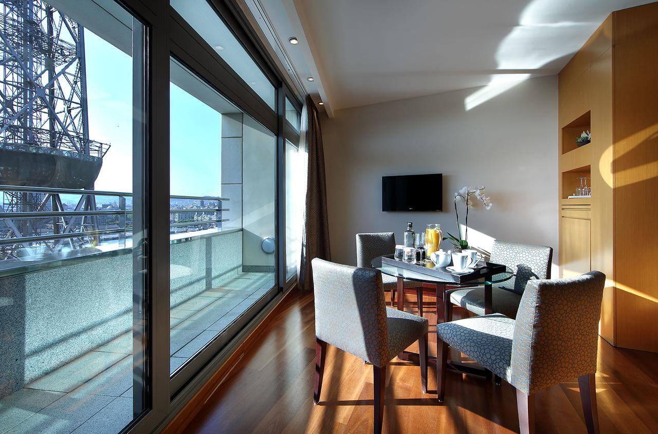 Eurostars Grand Marina Hotel Gl Barcelona Exterior photo The photo shows a modern and bright room that features large windows with panoramic views. Inside, there is a circular dining table surrounded by upholstered chairs. On the table, there are items that suggest a meal or refreshments, such as drinks an