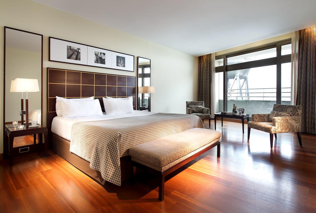 Eurostars Grand Marina Hotel Gl Barcelona Exterior photo The photo shows a modern hotel room featuring a large bed with a decorative headboard. There are two bedside lamps on either side of the bed, and behind the bed, there are framed pictures hanging on the wall. A bench sits at the foot of the bed, and 