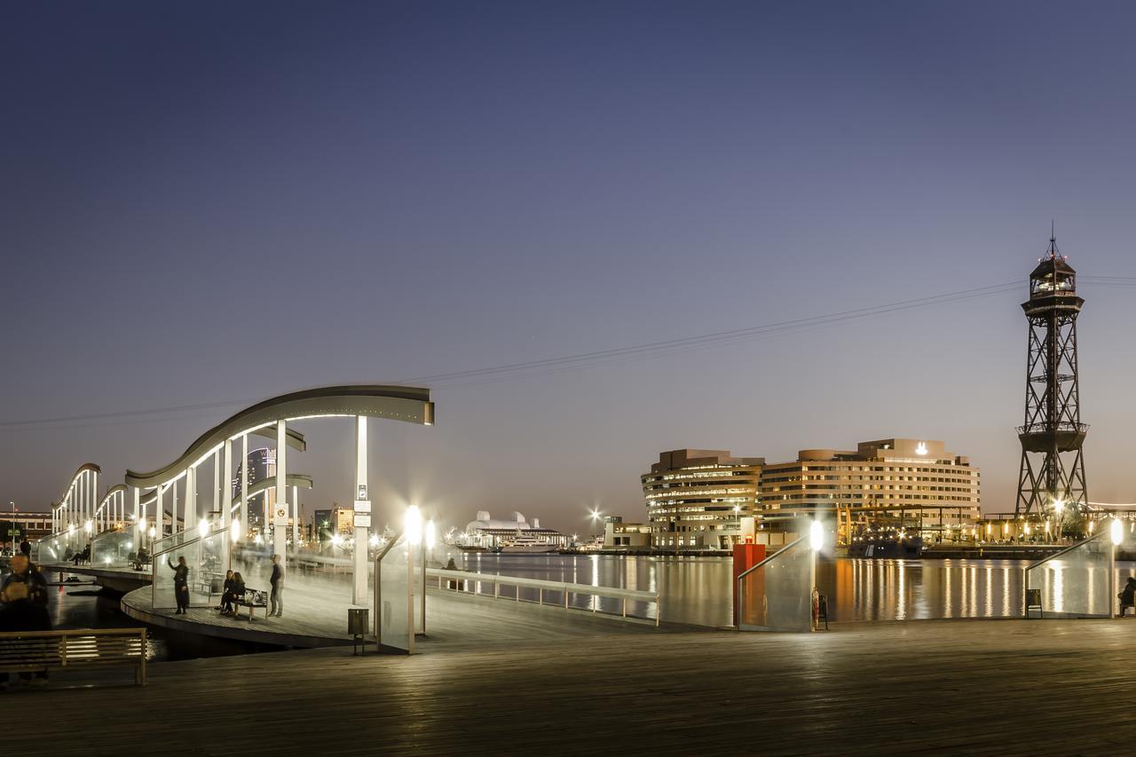Eurostars Grand Marina Hotel Gl Barcelona Exterior photo The photo depicts a scenic waterfront view during twilight. On the left, there is a modern, curved pedestrian walkway that appears to be well-lit, inviting pedestrians. To the right, a tall structure resembling a lighthouse or observation tower stand